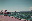 A coastal scene featuring boats, trees, and a serene ocean under a partly cloudy sky. The image has a slightly surreal color palette. blurred out low res