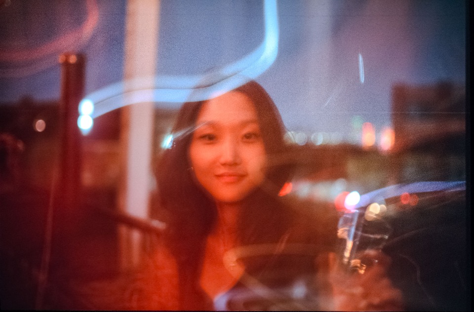 A soft, dreamlike portrait of a young woman, with streaks of light and blurred city lights in the background.