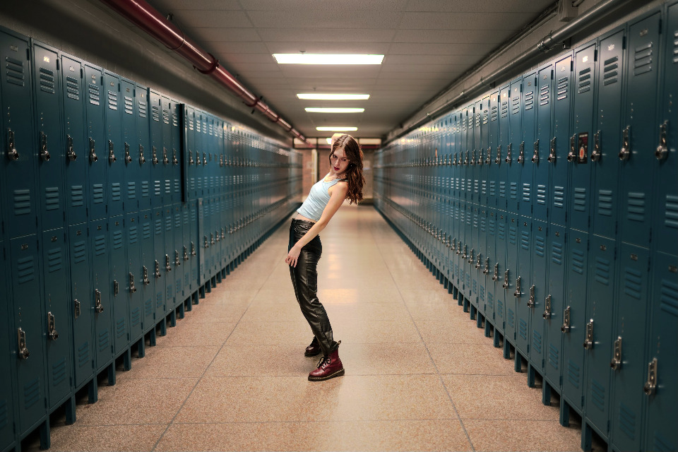 A woman poses in a school hallway, leaning back dramatically. The scene is framed by rows of teal lockers, leading the eye into the distance.